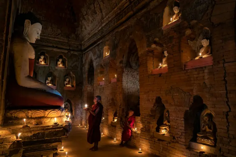 Monk holding candle in dimly lit Buddhist temple in Bagan, Myanmar, surrounded by Buddha statues.