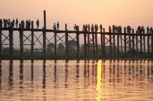 A serene sunset scene of people silhouetted on a wooden bridge with a reflection on the water.