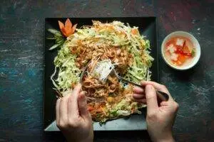 Top view of hands holding fork and spoon with noodles and fresh vegetables on plate.